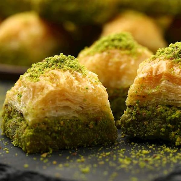 Close-up of three pieces of baklava with flaky pastry and green pistachio crumbles on a dark surface.
